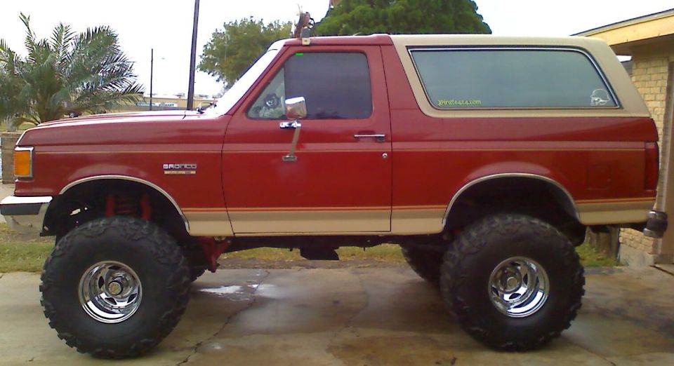 Side view of a red 1990 Ford Bronco with tan trim and large off-road tires