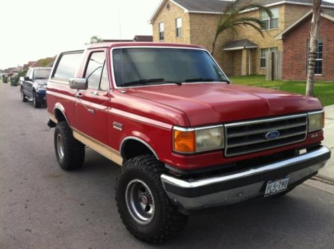 1990 Ford Bronco - This red 1990 Ford Bronco features a lifted suspension and off-road tires, parked on a street with houses in the background.