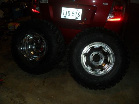 1990 Ford Bronco - Two aftermarket off-road tires and rims are shown in front of a red vehicle's rear.