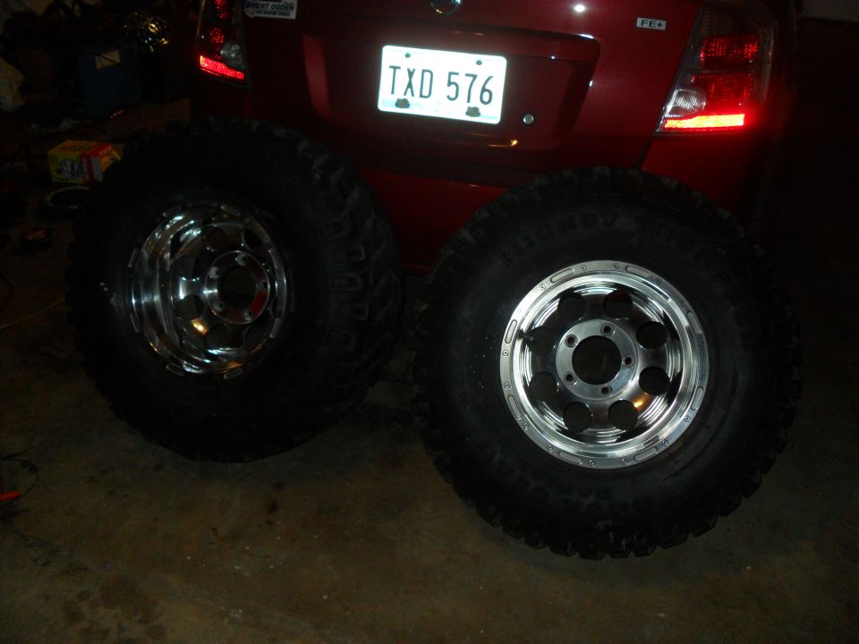 Two large off-road tires with chrome and silver rims are displayed in front of a red car's rear bumper and license plate
