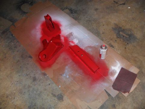 1990 Ford Bronco - Metal parts are being spray painted red on a cardboard surface, with a can of red spray paint and sandpaper nearby.