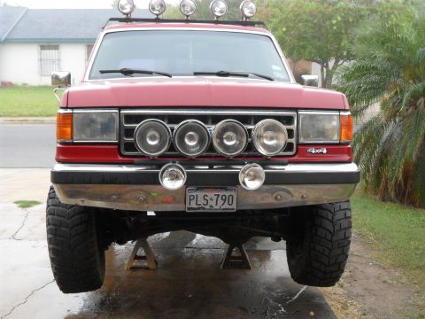 1990 Ford Bronco - This red Ford truck is on jack stands, showing its front end with multiple auxiliary lights and a 4x4 badge.