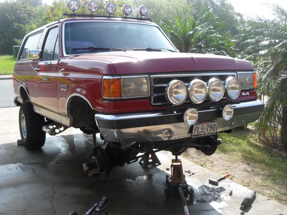 1990 Ford Bronco front end disassembled on jack stands, with auxiliary lights on roof and bumper