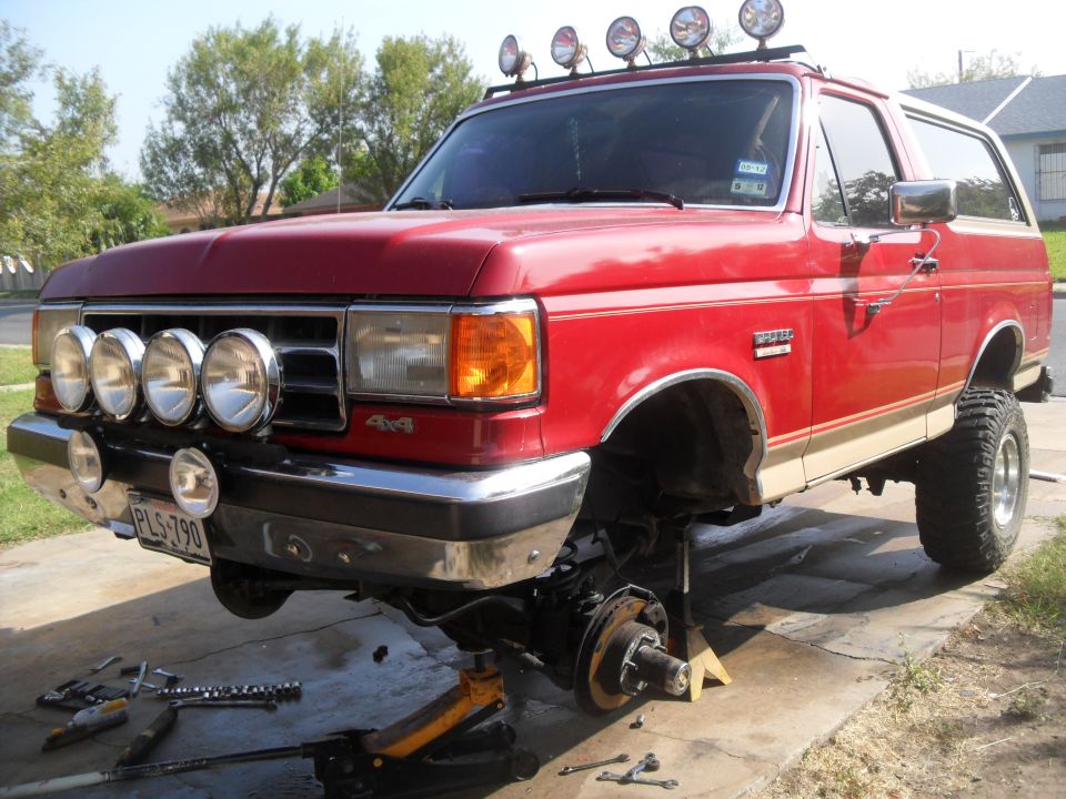 Red Ford Bronco on jack stands with front wheels off, tools scattered around