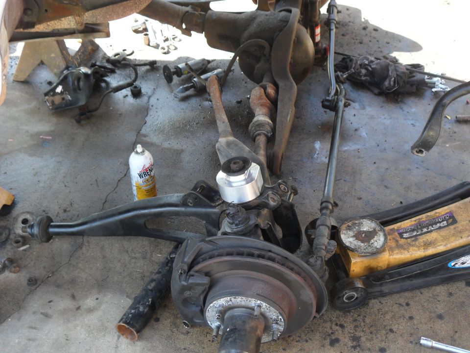 Vehicle suspension parts on the ground, including a brake rotor, control arm, and steering linkage. A can of spray lubricant is nearby