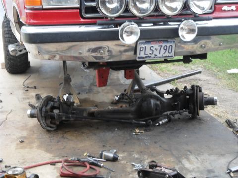 1990 Ford Bronco - The front axle assembly of a red truck has been removed and is resting on the ground, supported by jack stands.