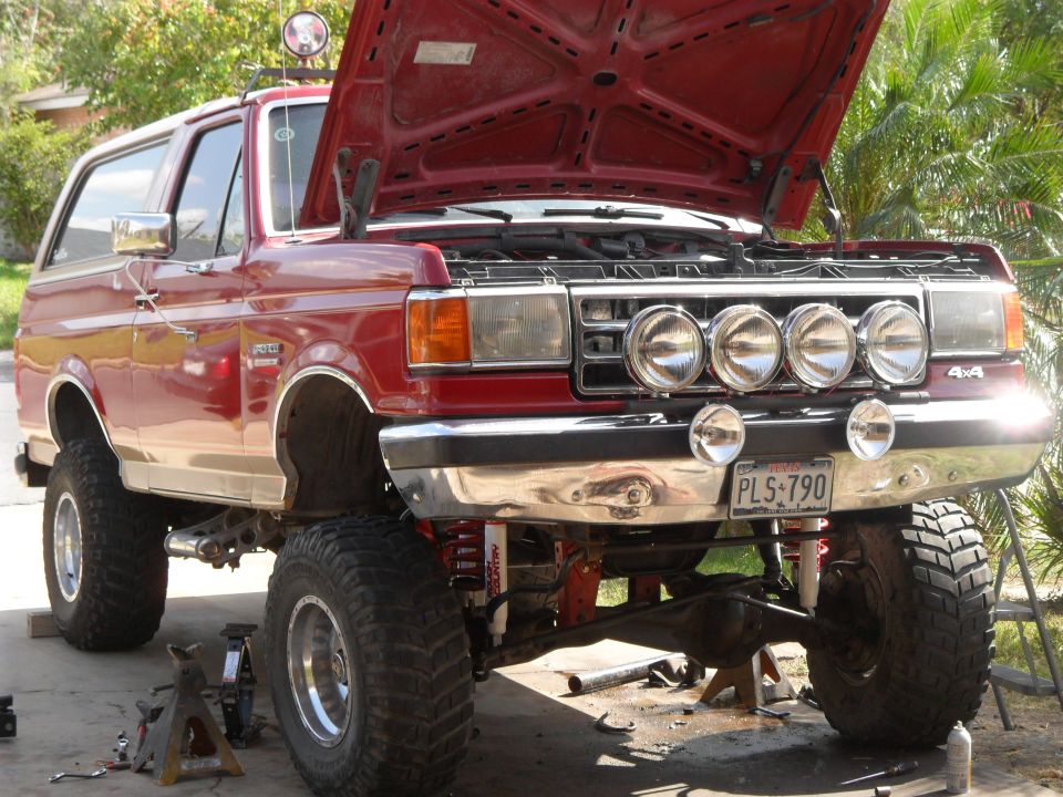 Red Ford Bronco with lifted suspension and hood open, four auxiliary lights mounted on the grille guard