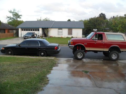1990 Ford Bronco - A lifted red Ford Bronco is parked next to a black sedan on a residential street.