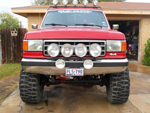 1990 Ford Bronco - This red SUV has a prominent grille with multiple auxiliary lights and a "Rough Country" decal on the windshield.