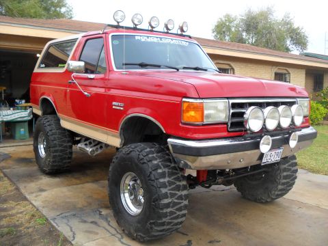 1990 Ford Bronco - A lifted red 1990 Ford Bronco with off-road lights and large tires is parked in a driveway.