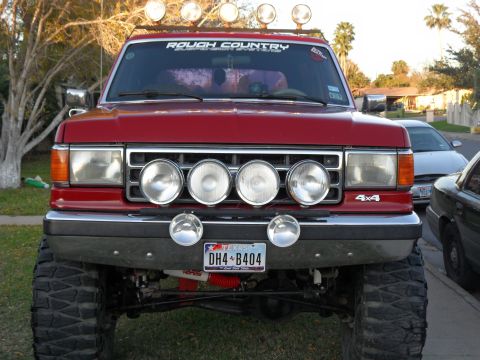1990 Ford Bronco - This red SUV features a prominent grille with four large driving lights and two smaller ones mounted below. A 'ROUGH COUNTRY' sticker adorn…