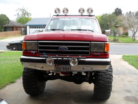 1990 Ford Bronco - This red Ford F-Series truck features a lifted suspension and large off-road tires, viewed from the front.