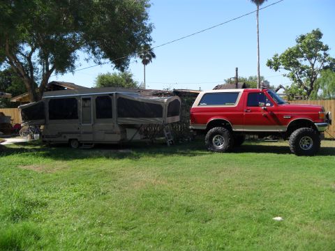 1990 Ford Bronco - A red 1990 Ford Bronco, lifted and equipped with off-road tires, is parked in a grassy yard next to a pop-up camper.