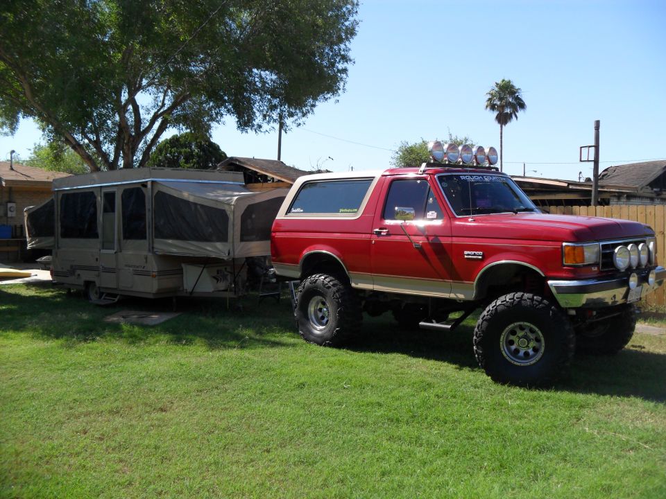 Red 1990 Ford Bronco with lifted suspension and off-road lights, towing a pop-up camper