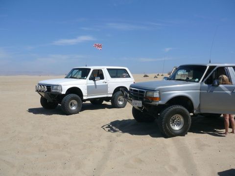 1988 Ford Bronco - Two Ford Broncos, one from 1988, are parked on a sandy beach with a checkered flag in the sky.