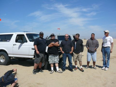 1988 Ford Bronco - A group of men pose for a photo with their vehicles in a sandy, outdoor location under a clear blue sky.