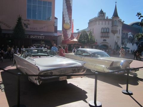 1991 Ford LTD Crown Victoria - Two classic cars are displayed outdoors in front of a themed building with people walking around.
