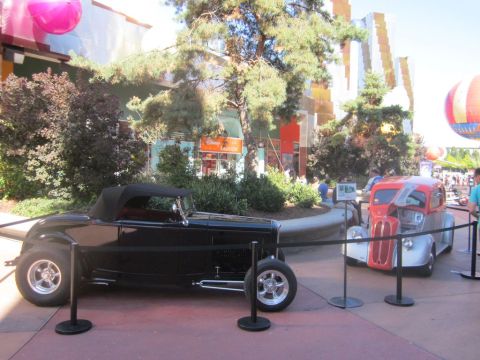 1991 Ford LTD Crown Victoria - Two classic hot rod cars are displayed outdoors with a building and trees in the background.