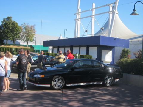 1991 Ford LTD Crown Victoria - A group of people stand around a black coupe parked in front of a blue and white building on a sunny day.