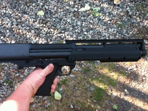2006 Ford E350 - Person holding a black bullpup-style shotgun by the pistol grip over a gravel driveway outdoors.