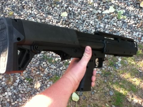 2006 Ford E350 - A black bullpup shotgun is held by a person's hand against a backdrop of gravel and grass.
