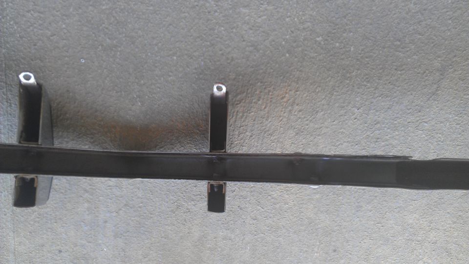 Black metal car part with two vertical supports, lying on a concrete surface. Supermotors.net logo in lower right