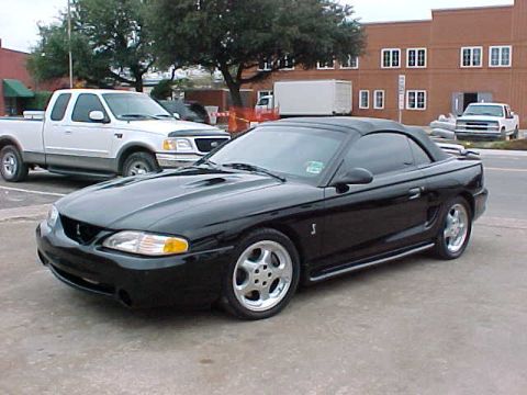 1995 Ford Mustang - Black 1995 Ford Mustang Cobra convertible is parked on pavement with a white pickup truck nearby.