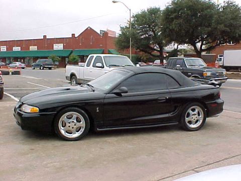 1995 Ford Mustang - A black convertible sports car is parked in a lot with other vehicles and buildings in the background.