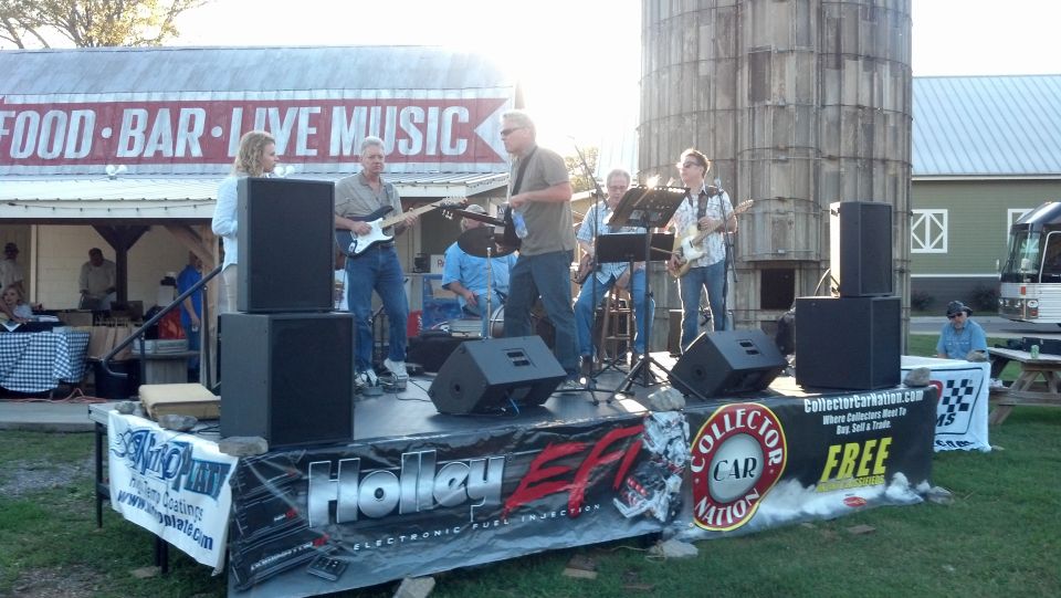 Band performing on an outdoor stage with speakers and banners, a silo in the background
