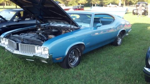 2007 Ford Mustang - A blue 1970s coupe, possibly an Oldsmobile 442, is displayed with its hood open at an outdoor car show.