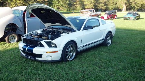2007 Ford Mustang - A white 2007 Ford Mustang GT500 with blue racing stripes is displayed with its hood open at an outdoor car show.
