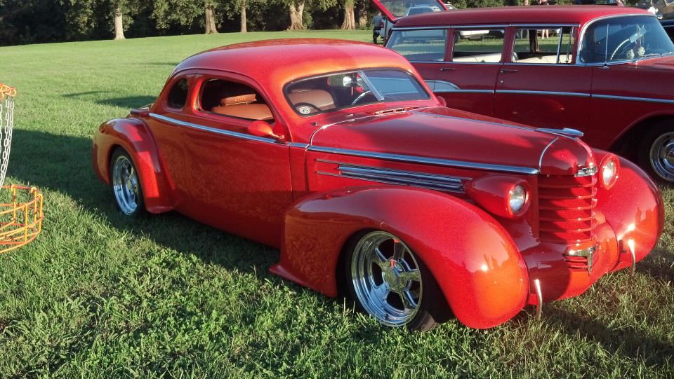 Red coupe hot rod with chrome wheels and custom grille, parked on grass next to a red station wagon