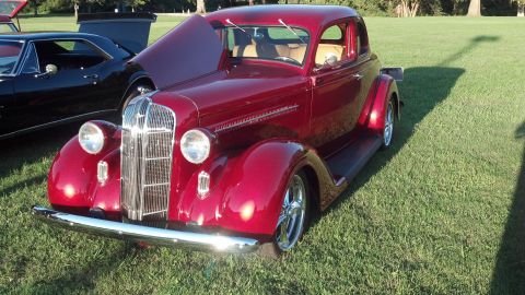 2007 Ford Mustang - A maroon 1930s coupe hot rod is parked on a grassy field next to another vehicle.