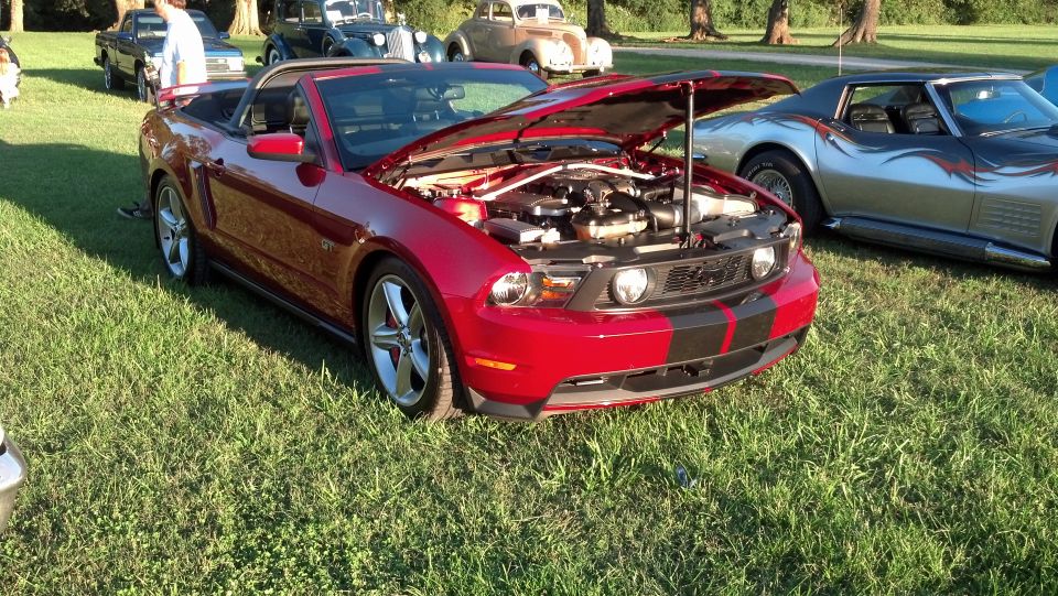 Red convertible with black racing stripes, hood open, parked on grass