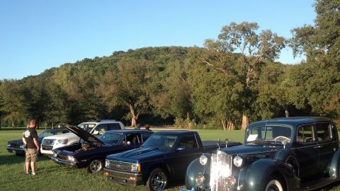 2007 Ford Mustang - A collection of vintage vehicles are displayed outdoors on a sunny day, with a lush green hill providing a scenic backdrop.
