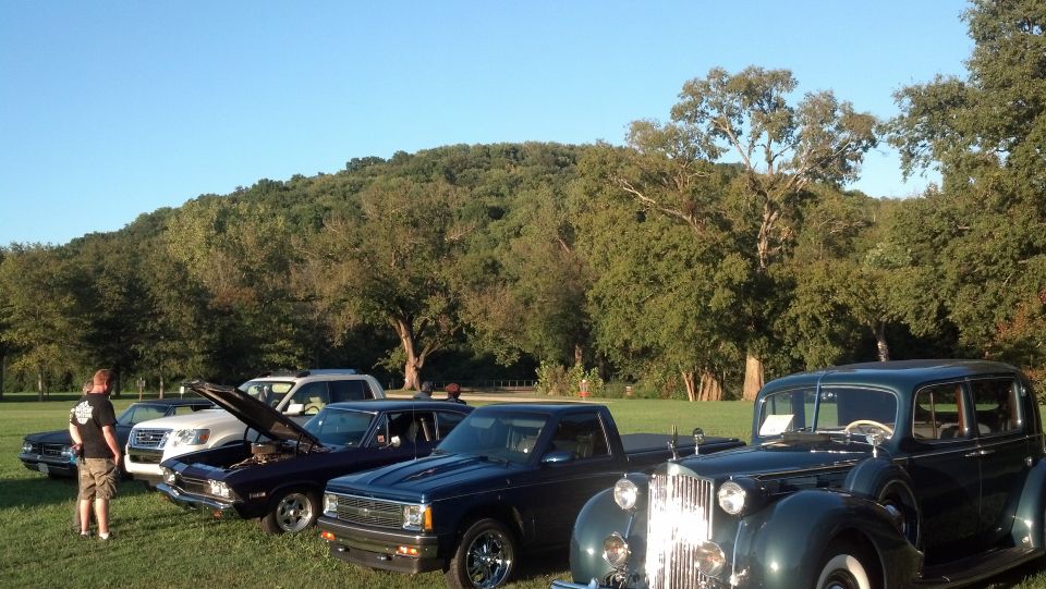 Several classic cars and a pickup truck parked on grass with a wooded hill in the background