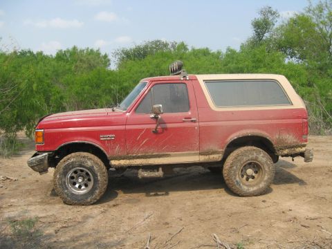 1990 Ford Bronco - This muddy red Ford Bronco is seen from the side, with a tan top and off-road tires.