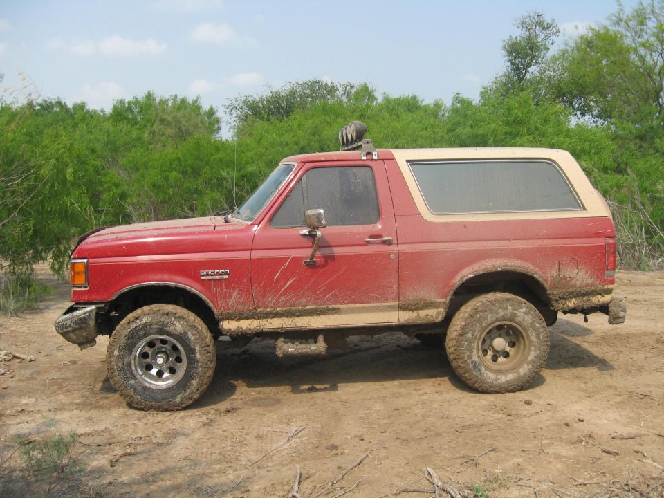 Red Ford Bronco with a tan top, covered in mud, parked on a dirt road with brush in the background