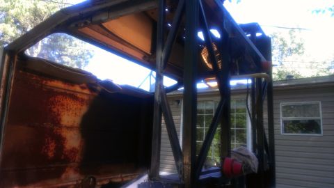 1978 Ford Bronco - The interior of a heavily rusted vehicle shows a damaged frame and roll cage structure against a backdrop of trees and a house.