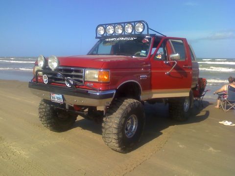 1990 Ford Bronco - A red Ford Bronco with large tires and extra lights sits on the sand with the ocean in the background.