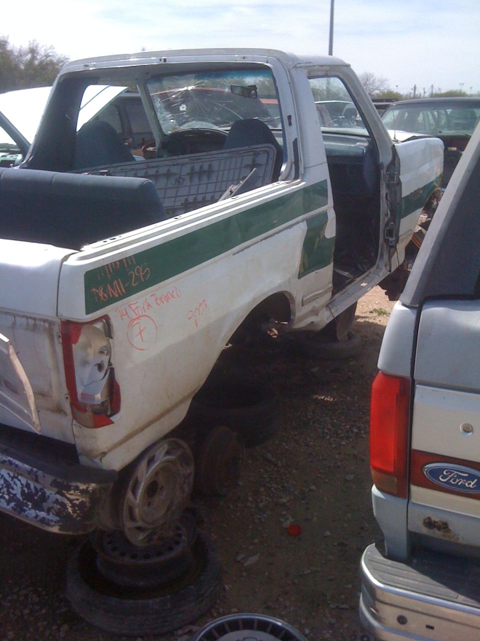 White pickup truck with green stripe, missing doors and wheels, parked in junkyard. Another Ford truck is partially visible to the right