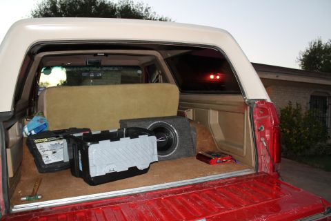 1990 Ford Bronco - The open bed of a red pickup truck contains a subwoofer, toolboxes, and other items.