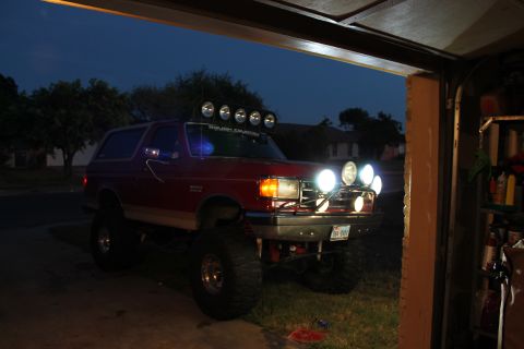 1990 Ford Bronco - A red 1990 Ford Bronco is parked outside a garage, equipped with multiple auxiliary lights.