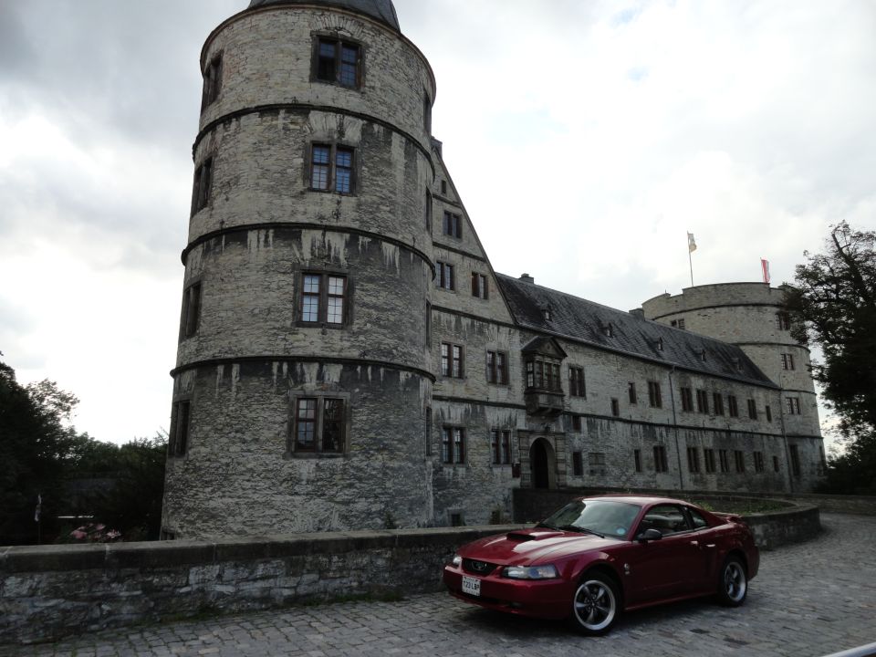 Red Ford Mustang parked on cobblestone in front of a stone castle