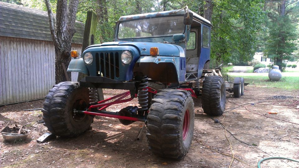 blue Jeep CJ with huge tires and red suspension parts, parked outside near a shed