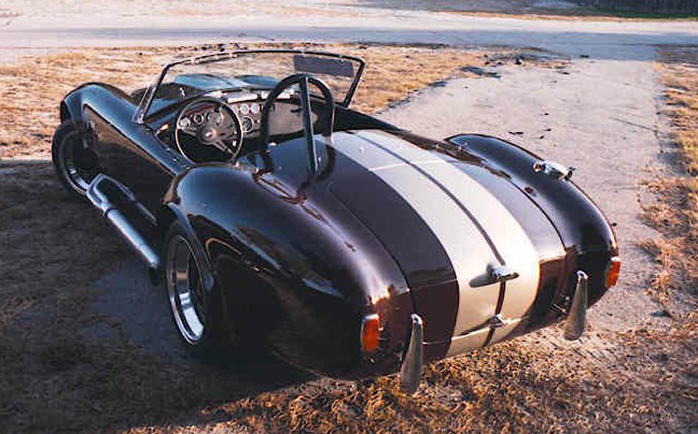 Black Cobra roadster with white stripes, rear 3/4 view, side pipes, on dirt lot
