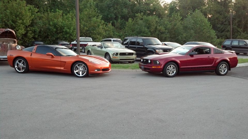 Orange Corvette and red 2007 Ford Mustang parked, with other vehicles and trees in background