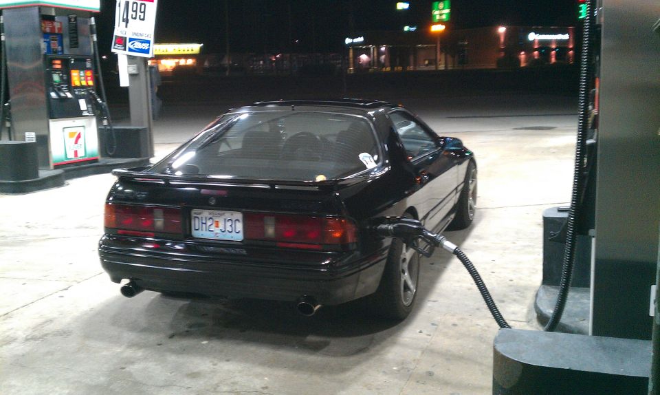 Black 1988 Mazda RX-7 with a gas nozzle in the fuel door, parked at a gas station at night