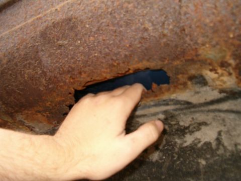 1990 Ford Bronco - Close-up of a heavily rusted inner fender well with a hand pointing to a rust-through hole eaten along the seam where the panel meets the f…