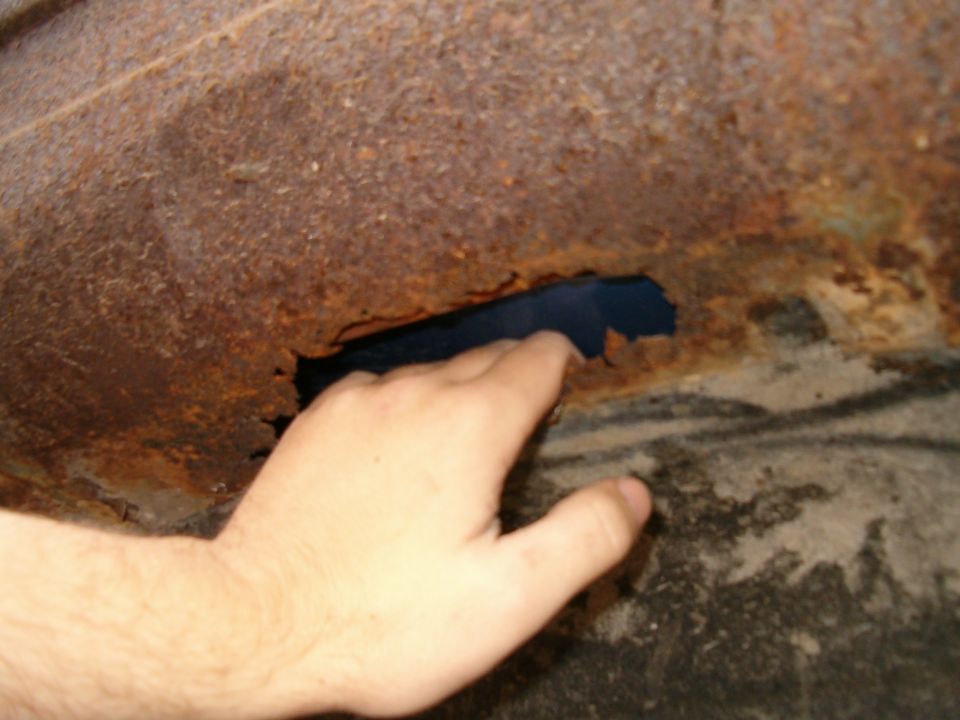 Hand pointing to rust-through hole in corroded inner fender well seam